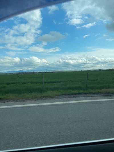 The image shows a view from a vehicle, likely a car, traveling on a road in Vodenichane, Bulgaria. To the right, the gray asphalt road is visible with a white line marking the edge. A green field stretches along the other side of the road with scattered posts or markers extending into the distance. The sky fills the upper left portion of the frame and is filled with blue sky and white clouds. A partial view of the vehicle's window frame is seen on the left and right edges.