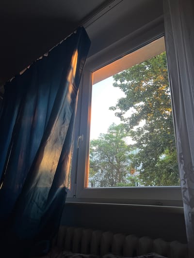 A dark blue curtain is pulled to the left, allowing sunlight to illuminate a portion of its fabric. To the right of the curtain is a window with a white frame. Through the window, trees with green foliage are visible, with a hint of buildings in the background. The sunlight filtering through the trees suggests it is either morning or late afternoon. Below the window, a radiator with white fins is visible, and a portion of a bed with dark bedding is in the foreground. The scene appears to be an interior view from a room in Skawina, Poland, looking out onto an urban or suburban environment.