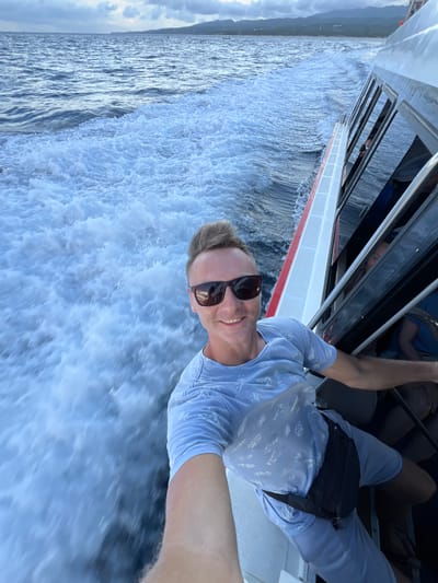 A cheerful man takes a selfie while riding on a fast-moving boat in Jungutbatu, Indonesia. The image is captured from a slightly low angle, showing the man smiling broadly at the camera, his face partially obscured by dark sunglasses. He has light brown hair and is dressed in a light blue t-shirt with a subtle white patterned print, light blue shorts, and a black fanny pack worn across his body. His arms are visible, suggesting he is holding on or positioning himself for the shot.
The boat is creating a dramatic, foamy white wake that churns intensely beside and behind it, indicating rapid movement across the water. The boat's side is visible, featuring a clean white hull with a thin red stripe and a dark-windowed cabin structure. Beyond the immediate turbulence, the ocean stretches out in a calmer, silvery-blue tone towards a hazy, distant shoreline or mountain range under a bright, partly cloudy sky. The overall scene conveys a sense of exhilaration and enjoyment during a daytime excursion. There is no visible text.