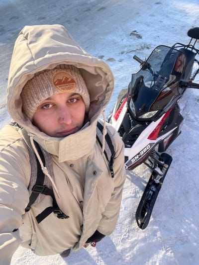 An overhead-angle selfie taken in Васильево, Russia, depicts a male individual standing on snow and ice next to a snowmobile. The man wears a light beige hooded winter coat, a beige knit beanie with an embroidered "MOUNTAIN PATHWAYS" patch, and a dark-strapped backpack visible over his shoulders. He has short facial hair and a small piercing below his lower lip. To his right, a black, white, and red snowmobile is parked, displaying "SN-380" on its side, with its handlebars, windshield, seat, and front skis visible. The ground consists of uneven snow and patches of ice under bright natural light.