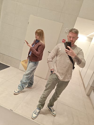 A mirror selfie captures two people in a public restroom. A man in a beige sweater and olive pants holds his phone to take the picture. He sports a watch and visible tattoos. Beside him stands a woman in a brown quilted jacket, blue jeans, and a light yellow tote bag. She holds a phone and looks at it. The restroom has a tiled floor, light-colored walls, and visible doors along the right.