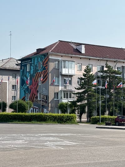 The image shows a mural on the side of a multi-story building in Belovo, Russia. The mural depicts a military theme, with images of soldiers, a red star, and the Order of St. George ribbon. The building has multiple windows and balconies. Russian flags are flying on poles in front of the building, as well as on the mural itself. Trees and bushes are visible in the foreground, and an antenna is mounted on a nearby building. In the background, a red car is parked. The street in front of the building has chalk drawings on it. The sky is clear and blue.