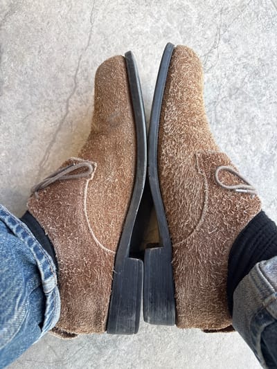 A top-down forensic view displays a person's lower extremities, specifically two worn, light brown, textured suede or nubuck lace-up shoes. The shoes are positioned parallel, their dark-colored inner soles and heels touching or nearly touching. Light brown laces are visible on each shoe. The individual wears dark blue denim jeans, cuffed at the ankle, and black socks, which are visible between the jean cuffs and the shoe uppers. The footwear rests on a light gray, subtly textured floor, consistent with concrete or large format tiles, exhibiting minor surface variations. No specific visual information within the image allows for confirmation of the stated geographic location in Зеленогорское сельское поселение, Ukraine.