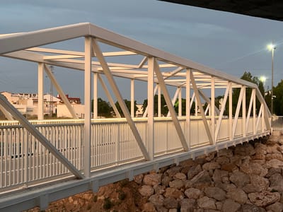 This image displays a pedestrian truss bridge situated in Alcàsser, Spain, observed during either dusk or dawn. The bridge is constructed from off-white or light gray painted metal, featuring a series of interconnected rectangular and triangular hollow sections forming its upper structure. Along both sides, a matching light-colored vertical slat railing is installed. Integrated linear lighting runs along the top of the railings, illuminating the bridge's pathway. The bridge spans over a terrain characterized by numerous large, irregularly shaped brown and gray rocks, interspersed with reddish-brown soil and sparse green vegetation. In the background, under a soft blue-gray, overcast sky, a line of distant buildings with light-colored facades and some reddish roofs is visible. Scattered green trees are present beyond these structures. On the right side of the frame, several streetlights are illuminated, emitting bright white light and producing distinct lens flares. No individuals are present on the bridge or in the immediate vicinity, and no specific actions or interactions are depicted.