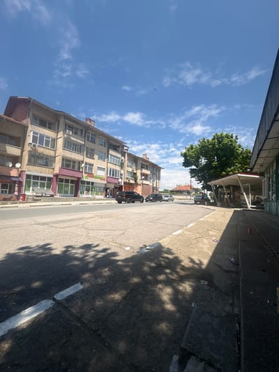 The image depicts a street scene in the village of Tenevo, Bulgaria, under a clear to partly cloudy blue sky. The sun is high, casting distinct shadows of trees and structures across the road and sidewalk, suggesting a mid-day setting.
On the left side of the asphalt road, a multi-story building with a light-colored, somewhat weathered facade dominates. This building features numerous windows, some with enclosed balconies, and several satellite dishes affixed to its exterior. The ground floor houses multiple commercial establishments; one shop displays "Млечен магазин" (Dairy store) and another "Храни" (Food), both with large glass storefronts, some painted green or burgundy. A black SUV is parked on the road in front of these shops.
The road itself is paved with asphalt, marked with a broken white line along its left edge. Tree shadows are prominent across its surface.
On the right side of the image, a concrete sidewalk is visible, appearing uneven and scattered with litter. A prominent green, leafy tree provides shade over a bus stop or public shelter. This shelter has a white roof with red trim and a glass enclosure. Several individuals are gathered near and under this tree and shelter; one person is seated within the shelter, and others are standing nearby. Further right, a partial view of a single-story building with a weathered, possibly green-painted facade is present.
In the background, beyond the main intersection, additional residential buildings with tiled roofs are visible, along with other parked and moving vehicles. A small bird is discernible in the upper left portion of the sky.