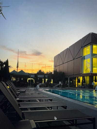 The image, which appears to be a photograph of a digital screen as evidenced by visible pixel grid patterns and a mouse pointer icon in the lower right quadrant, depicts an outdoor swimming pool facility at dusk or sunset. A row of dark, unoccupied lounge chairs is arranged along the left edge of a rectangular pool, which contains blue water with visible lane dividers and entry handrails. To the right of the pool, a modern, multi-story building with a brown, geometrically paneled facade exhibits brightly illuminated yellow windows on its ground and upper floors. In the mid-ground, to the left of the pool, a tall flagpole displays a partially unfurled Canadian flag. Nearby are several dark green-canopied structures, possibly tents or large umbrellas. Various dark trees and and shrubs are visible around the perimeter of the pool area. A light-colored fence and distant silhouetted structures are present in the background against a sky transitioning from orange and yellow near the horizon to pale blue above. No individuals are present in the scene. The presence of the Canadian flag indicates the location is in Canada, situated within Electoral Area A, British Columbia.