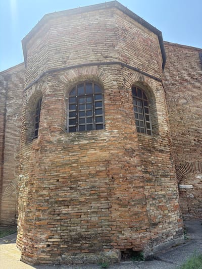 The image shows an exterior view of an octagonal brick structure with arched windows. The bricks are weathered and show variations in color, ranging from reddish-brown to lighter tan. Two arched windows are visible on the curved section of the structure, each with wooden frames and multi-paned glass. The building appears to be old and possibly historic, given the condition of the bricks and the architectural style. The sky is clear and bright, with some sunlight casting shadows. The ground around the structure is paved with asphalt and there is some sparse vegetation growing at the base of the wall. The building is identified as the Church of San Michele Arcangelo in Santarcangelo di Romagna, Italy.