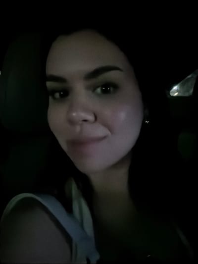 The image presents a close-up, chest-up portrait of an adult female, centrally framed. The subject has dark hair, dark eyebrows, and dark eyes, looking directly into the camera with a subtle, closed-mouth smile. Her complexion is fair, with a slight sheen on the cheeks and forehead, suggesting frontal or slightly off-center illumination. A light-colored, likely white, strap from a garment is visible over her left shoulder. The background is predominantly dark, indicative of a low-light environment. On the left side, a dark, textured surface, consistent with a vehicle's interior upholstery, is discernible. On the right, a small, bright, reflective anomaly is visible against the dark background. The scene suggests a selfie taken inside a vehicle or a confined, dimly lit space. The specific location within the city of Veracruz, Mexico, cannot be determined from the visual evidence provided.