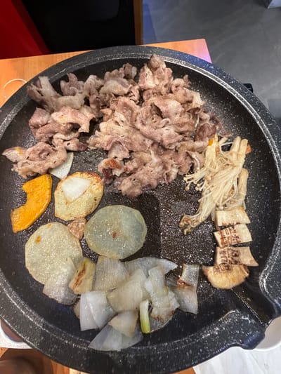 A top-down view captures a variety of food cooking on a black, speckled griddle. On the left, a pile of thinly sliced, light brown meat (possibly beef or pork) is sizzling. To the top-middle, a cluster of white enoki mushrooms is lightly cooking alongside several rectangular, light-brown pieces of food, which appear to be some kind of cake or tofu. These rectangular pieces have dark, unidentifiable characters imprinted on their surface and show some browning.

In the bottom-middle, several thin, round slices of potato are visible, some with lightly browned edges, indicating they are being grilled. A single crescent-shaped piece of orange pumpkin is also cooking nearby. To the right, irregularly cut chunks of a white vegetable, likely daikon radish or onion, are spread out, appearing somewhat translucent. The griddle itself is a dark, non-stick surface, suggesting an indoor dining setting where food is cooked at the table. The overall scene depicts a communal cooking experience, typical of East Asian cuisine.