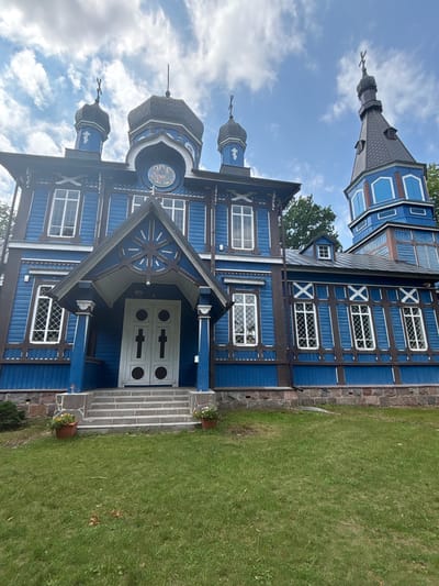 The image shows a bright blue wooden church with dark brown trim, located in Puchły, Poland. The church features multiple domes topped with crosses, and a prominent bell tower to the right. The facade of the church is adorned with decorative wooden carvings and features large, white-framed windows. A covered entrance with double doors, also painted white, is approached by a set of stone steps. Two potted plants with yellow and white flowers sit on either side of the steps. The church is surrounded by a green lawn, and the sky above is partly cloudy. The perspective of the photo is from a low angle, looking up at the structure.