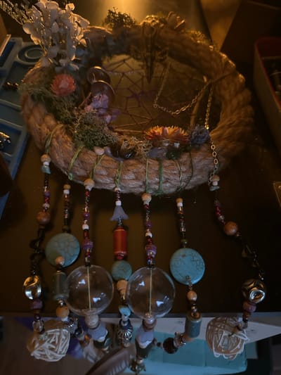 The image shows a close-up, overhead view of a handmade dreamcatcher-like object, likely a wind chime or wall decoration. The object is circular and appears to be constructed from thick, braided rope. It is adorned with various natural elements such as dried flowers, moss, and what look like small pinecones or seed pods. Dangling from the bottom of the circular frame are several strands, each made of numerous beads of different shapes, sizes, and colors, including turquoise, wood, glass, and metallic elements. Some strands also feature larger decorative elements like clear glass orbs, a cylindrical red bead, and intricate woven natural fiber spheres. The background is dark and appears to be a tabletop or shelf. The lighting is warm and casts shadows. There is no discernible location within McMasterville, Canada evident in the image.
