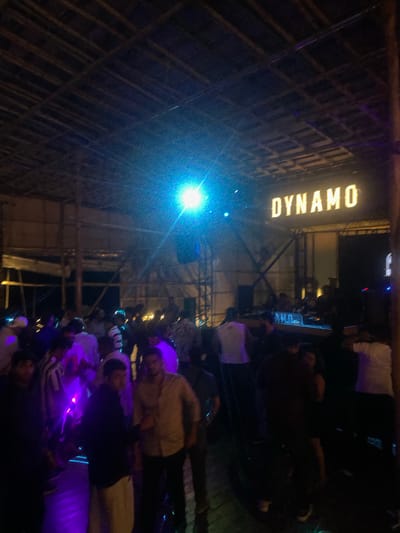 The image depicts a nighttime scene at a venue named "DYNAMO" in Vagator, India. A crowd of people is gathered, likely at a club or outdoor event. The environment appears to be somewhat rustic, with a thatched or bamboo roof structure visible overhead. Bright, colored lights, including a prominent blue light and purple lighting effects, illuminate the scene, casting various hues on the people and surroundings. In the background, a DJ booth or stage area is discernible with some equipment. The individuals in the foreground and midground are dressed in casual attire, suggesting a relaxed atmosphere. Some people are standing, while others appear to be dancing or socializing. The overall impression is one of a lively, nighttime gathering at a popular entertainment spot in Vagator.