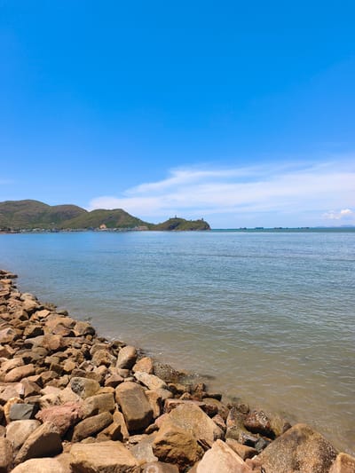 This is an eye-level, full shot of a rocky shoreline on a bright, clear day. The foreground is dominated by a dense arrangement of various-sized, rough-hewn rocks, ranging in color from light brown to reddish-brown, with some showing white mineral deposits. The rocks form a natural barrier between the land and the water. The water itself is a muted, greenish-blue, with a gentle ripple pattern visible. In the mid-ground, a town with low-rise buildings and a church-like structure is nestled at the base of green, rolling hills. A small island with a lighthouse-like structure and another building stands prominently on the right side of the frame, further out in the sea. Several cargo ships are visible as small silhouettes on the horizon. The sky above is a vibrant, clear blue, with a few wispy white clouds scattered across the upper portion. This scene is likely located near the coast of An Nhơn, Vietnam, given the geographical features depicted.