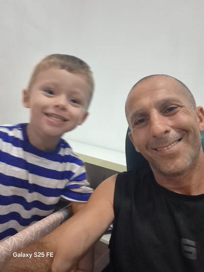 An indoor selfie shows two individuals, likely father and son, smiling at the camera. The child, positioned on the left, wears a blue and white striped t-shirt and has light brown hair. The man, on the right, is bald, has a light beard, and wears a black sleeveless shirt. They appear to be in a home environment, with a blurred background that includes a desk and wall. The text "Galaxy S25 FE" is visible in the lower left corner, indicating the device used to take the photo. The scene is brightly lit, suggesting artificial indoor lighting.