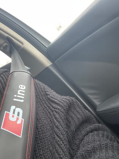 The image shows a close-up view inside a vehicle, specifically focusing on the seatbelt. The seatbelt has a black leather cover with red stitching and the "S line" logo in white and red. A portion of a dark grey knitted garment is visible behind the seatbelt cover. The vehicle's interior, including a perforated grey panel and part of the roof, is also visible. The scene appears to be taken from a slightly upward angle. The location information is not discernible from the image.
