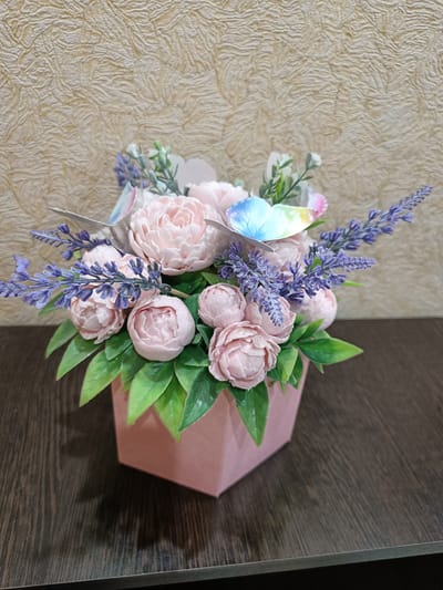 This media file shows a close-up of a decorative floral arrangement in a pink hexagonal vase. The arrangement features several light pink peony-like flowers and sprigs of artificial lavender. Two iridescent butterflies are placed amongst the flowers, one on the left and one in the center, with the latter displaying blue and rainbow hues. Green artificial leaves fill the spaces between the flowers and frame the vase. The arrangement is placed on a dark brown wooden surface, possibly a table, which has a noticeable wood grain. The background is a textured, light beige wallpaper with a raised, wavy pattern. The lighting appears to be artificial and is casting a soft shadow from the vase onto the surface. There is no text visible in the image. The overall impression is that of a whimsical and delicate decorative piece. The setting suggests an indoor environment, likely a home or a craft display.