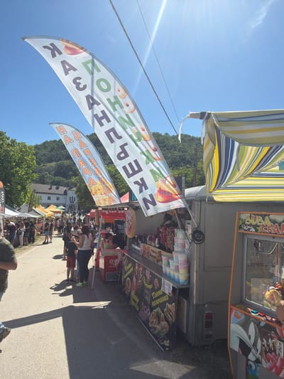 The image shows an outdoor market or festival scene in Ustrem, Bulgaria, based on the Cyrillic signage. A food vendor's stall is visible, featuring a striped awning and banners advertising "Казанлъшки Kъфки" (Kazanlak Donuts) and "Пържени Палачинки" (Fried Pancakes/Crepes). People are walking along a road. A crane game is attached to the stall. The backdrop includes greenery and building tops, indicating a setting with hills or a raised terrain. The sky is blue with sunlight.