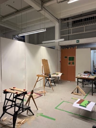 An art studio features an easel facing a white wall. A wooden backdrop with green-painted squares stands nearby. A worktable holds art supplies. A black workbench is positioned in the foreground. The floor has green tape markings. The studio's ceiling reveals industrial lighting and beams, enhancing the space's functional and raw aesthetic. A ladder cage is visible above a doorway in the top left corner.