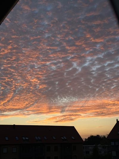 The image captures a sky filled with altocumulus clouds, tinged with hues of orange and pink from a sunset or sunrise, as they are illuminated by the low-lying sun. Below the sky, the silhouettes of buildings are visible, including a residential building with a dark red tiled roof and multiple dormer windows. The buildings appear to be located in a city, with a faint outline of trees and other structures in the background. The overall scene suggests an urban environment at dusk or dawn. Based on the architectural style of the buildings, particularly the red tile roofs and dormer windows, this scene is consistent with residential areas found in Roskilde, Denmark.