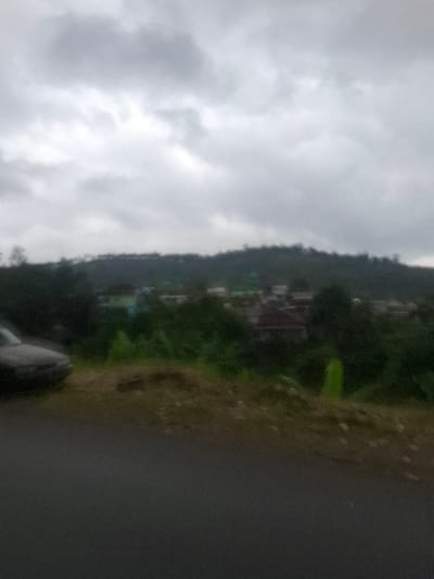 Overcast sky dominates, transitioning to a verdant hillside with buildings nestled among trees. The landscape suggests a settlement perched on an incline. A paved road, slightly blurred, occupies the foreground, implying the image was taken from it. The day appears gloomy and damp.