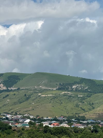 The image depicts a view of the city of Ust-Dzheguta, Russia, nestled at the foot of verdant, rolling hills. The sky is filled with dramatic, cloudy formations, suggesting an overcast day. In the foreground, a dense cluster of residential buildings with various roof colors, including red, green, and white, forms the urban landscape. These houses are interspersed with trees. The middle ground is dominated by the undulating green slopes of the hills, marked by lighter patches of dry grass and scattered trees, and crisscrossed by visible pathways or eroded tracks. The background features higher, more rugged mountain terrain, with exposed rocky areas contrasting with the green vegetation. There are no people or specific actions visible in the image.