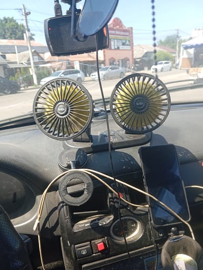 This media file shows the interior of a car, looking out through the windshield. The focus is on the dashboard, which is cluttered with various objects. Prominently displayed are two black and yellow car fans, positioned side-by-side. Above them, a dashcam is mounted. A smartphone is also visible, secured in a holder on the dashboard. Several wires and cables are draped across the dash, connecting various electronic devices.
In the background, visible through the windshield, is a street scene under a clear, sunny sky, suggesting daytime. Buildings, trees, and other vehicles are discernible, indicating an urban or suburban environment. The overall impression is of a functional but somewhat messy car interior, likely in use during a road trip or daily commute. The presence of the fans and the sunny weather suggest a warm day. No people are visible in the media file.
There is some visible text on the dashcam and possibly on the phone screen, but it is not clearly legible. The text on the fans is also too small to read. The overall mood is mundane and everyday, capturing a slice of life from inside a vehicle. The location context provided is Vasylkiv, Ukraine.