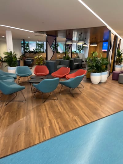 This image captures a bright and modern interior space, likely a waiting area or lounge. The perspective is slightly angled, looking across the room. The floor is a prominent feature, distinctly divided into two sections by a diagonal line: the left side features a light brown wood-patterned laminate or vinyl, while the right side is a uniform, light blue or teal color.
Numerous contemporary chairs are arranged in rows across the wood-patterned floor. These chairs have molded seats and sleek metal legs, with some chairs colored a vibrant coral/orange and others a cool teal/light blue. No people are visible, suggesting the area is currently unoccupied.
In the background, the space continues with additional seating arrangements, including darker upholstered armchairs or sofas. Large green plants in stylish white planters, some adorned with light blue stripes, are strategically placed throughout the room, enhancing the aesthetic.
The walls are part of a modern design, with a section of wood paneling visible in the mid-ground. Mounted on this wooden wall is a blue screen or monitor displaying white text; "24/7" is clearly legible, indicating a service or information display. Further back, the architectural details include geometric patterns and mirrored surfaces that reflect parts of the room. A long, linear light fixture spans the ceiling, contributing to the bright and evenly lit environment.
The overall impression is one of a clean, well-maintained, and thoughtfully designed contemporary space. The location cues, such as the modern design and "24/7" display, are consistent with a public building, potentially a university common area, office reception, or healthcare facility in Kelvin Grove, Australia. The time of day cannot be determined due to the indoor setting and artificial lighting.