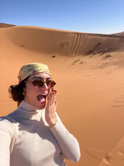 A young adult female is captured in a close-up selfie in a desert environment near Rissani, Morocco. She is positioned slightly off-center to the left, looking directly at the camera with her mouth wide open, tongue partially visible, and her left hand raised with fingers extended next to her mouth, conveying an expression of excitement or surprise. She wears a light beige ribbed turtleneck sweater, a patterned headscarf featuring green, yellow, pink, and brown hues, and oval-shaped brown sunglasses. Her left wrist displays a gold bracelet, and her fingernails are painted red. The background consists of large, undulating orange-brown sand dunes under a clear blue sky, characteristic of the Erg Chebbi desert landscape. Subtle tracks and sparse, small patches of vegetation are visible on the distant dunes, with distinct shadow patterns cast by the sun.