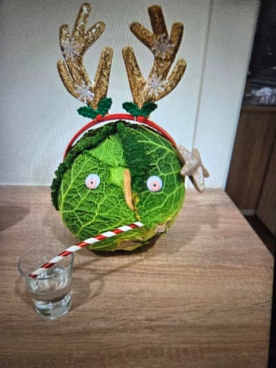The scene depicts a decorated head of green cabbage positioned on a light brown wooden surface, allegedly located in Blainville-sur-Orne, France. The cabbage is adorned with a red headband featuring two glittery gold reindeer antlers with white snowflake patterns and small green holly leaf embellishments. Two pink-rimmed googly eyes are affixed to the front of the cabbage, and a thin, brown/gold star ornament is attached to its upper right side. A red and white striped straw protrudes from the lower front of the cabbage, extending towards a small, clear glass tumbler partially filled with a transparent liquid, likely water. The glass rests on the wooden surface to the left of the cabbage. In the background, a light-colored wall with vertical textured lines is visible, with a darker brown vertical element, possibly furniture or a door frame, present on the far right. No people are visible in the image.