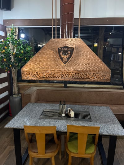 The image shows the interior of a restaurant. A decorative copper-colored hood with an emblem on it is prominently featured, possibly above a cooking area or grill. Next to the hood is a grey granite-topped table with a metal grilling area built into the top, and two wooden chairs. A small lemon tree is positioned near the base of the copper hood. Behind the hood is a window. Based on the presence of the word "ТАБИЕТ" on the copper hood, it can be assumed that the location is the "Tabiet" restaurant in Smilyan, Bulgaria. There are no people visible in the image.