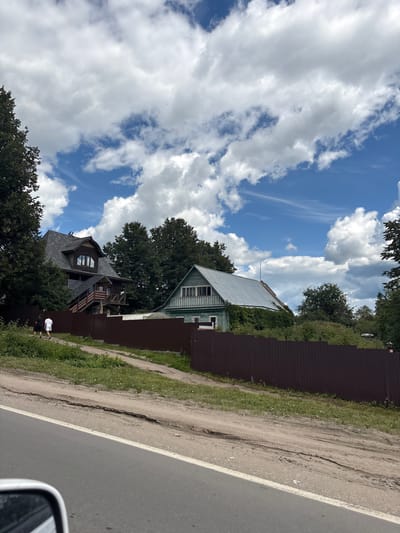 This image, taken in Pereslavl-Zalessky, Russia, depicts a scene from a car's perspective, showing a residential area by a road on a cloudy day. The road is paved with a white line marking the edge, and to its left is a dirt path with grass. A tall, dark brown corrugated metal fence runs along the path, separating it from the properties behind.
Behind the fence are two houses. The house closer to the foreground is dark with a complex, multi-gabled roof covered in dark shingles. It has a wooden, multi-level balcony structure attached. Further back and slightly to the right is a lighter-colored house with a light-colored metal roof and green siding. Both houses are surrounded by dense green trees.
In the distance, the sky is a vibrant blue filled with dramatic, fluffy white clouds. The overall impression is of a rural or suburban setting, possibly a dacha village, under a dynamic sky. The image is oriented vertically, making it appear as if the viewer is looking up or at a tilted perspective. The time of day appears to be daytime, given the bright sky and the way the light falls on the scene. There is no visible text in the image. A person in a white t-shirt and dark shorts is visible walking along the dirt path.