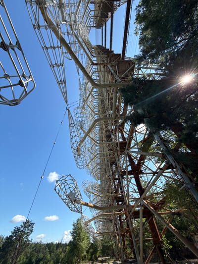 An extreme low-angle, wide shot captures the massive, rusting metal structure of the Duga radar array, a Cold War-era Soviet over-the-horizon radar system, under a clear blue sky. The colossal array dominates the frame, its intricate network of steel beams and wires stretching upwards and outwards. Partially obscuring the structure on the right side of the image are the dense green branches of coniferous trees, with sunlight glinting through them, creating lens flare. The environment appears overgrown, with more trees visible in the background, suggesting a natural, unmaintained setting within the Chernobyl Exclusion Zone. The sky is a vibrant, cloudless blue, providing a stark contrast to the weathered, industrial metal of the radar. The perspective is disorienting, with the ground and sky appearing inverted or at unusual angles, emphasizing the sheer scale and imposing nature of the structure. No people are visible in the scene.