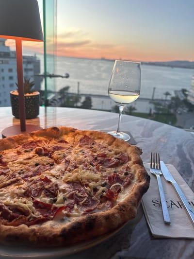 This image appears to be taken at a restaurant in İzmir, Turkey, based on the printed word on the napkin. The image shows a high-angle view of a pizza on a round table with a marble or similarly patterned surface. Beside the pizza, there is a wine glass filled with a light-colored liquid, possibly white wine. A set of silverware is on a napkin. In the background, through a window, there is a cityscape fading into a body of water. The sky suggests either a sunrise or sunset with orange and pink hues. In the lower left corner, a portion of a modern-style lamp is visible, as well as some indoor decorations.