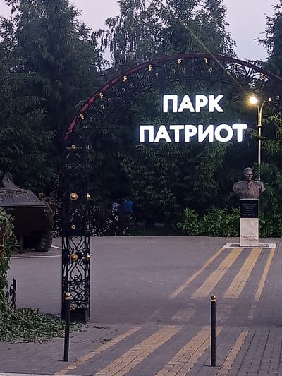 The image shows an outdoor scene in front of an entrance to "PARK PATRIOT" in Panino, Russia. A large, ornate black archway with gold accents spans the entrance. White, illuminated letters on the archway spell out "ПАРК" (Park) on the top line and "ПАТРИОТ" (Patriot) on the bottom line.
To the right of the entrance, a bust of a man in military attire is displayed on a pedestal. The background is filled with dense evergreen trees. A paved pathway with yellow striped crosswalk markings leads into the park.
On the left side of the image, partially visible, is the rear of a military vehicle, possibly an armored personnel carrier. A decorative black metal gate pillar stands in the foreground on the left. A few individuals, dressed in casual clothing, are visible in the middle ground, near the trees and behind the gate. The overall lighting suggests it is either early morning or late evening.