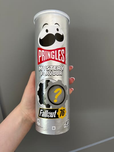 A person's hand is holding a Pringles can featuring a Fallout 76 collaboration. The Pringles can is silver with red and black accents. The iconic Pringles mascot is on the left side of the can, with a surprised expression. To the right of the mascot, "PRINGLES" is written in large red letters. Below that, "MYSTERY FLAVOUR" is written in smaller black text. In the center of the can is a circular graphic with a gear motif and a yellow question mark. To the right of this graphic, "Fallout 76" is written in a bold yellow font. There are small nutritional information details and a QR code on the right side of the can. The background is a plain gray wall, suggesting an indoor setting. The lighting is even and diffused, with no strong shadows. The hand holding the can appears to be a fair-skinned individual with white nail polish on their fingers. The image is taken from a slightly elevated angle, looking down at the can.