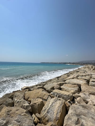 A wide-angle, high-angle shot depicts a clear sky over a coastal area. The sea, with its turquoise and blue hues, meets a shoreline lined with a breakwater made of large, light-colored rocks. The waves are breaking, creating white foam against the rocky structure. In the distance, a town with buildings and hills is visible along the coast. The scene is natural and serene, with no people or animals present. This landscape suggests a location within the Kissonerga Community in Cyprus, known for its coastal beauty and rocky shores.