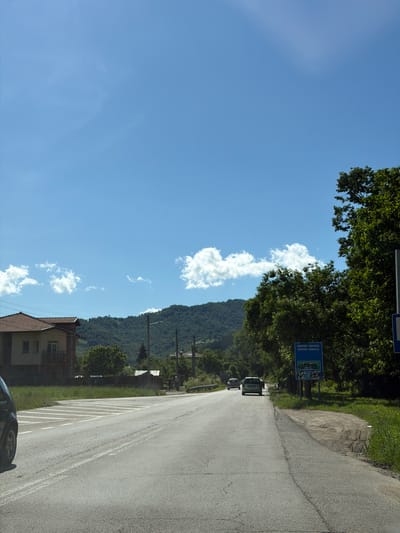 The image depicts a rural road scene in Beli Osam, Bulgaria, under a clear blue sky with scattered white clouds. A paved road, with visible white lane markings and some wear and tear, stretches into the distance. On the left side of the road, there is a building with a tiled roof and a balcony. Further back, a treelined hillside forms the backdrop. On the right side of the road, a dense cluster of green trees is prominent. Attached to a pole within the trees is a blue sign with white lettering that reads "ШИПКООВО-РИБАРИТСА SHIPKOVO-RIBARTSI" indicating a place name. Below this sign, there is another sign with green and white text and a graphic. A few cars are visible on the road, with one car in the foreground on the left, partially cropped, and two cars further down the road, moving away from the viewer. The environment appears to be a warm, sunny day.