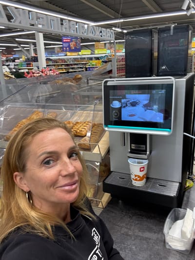 A woman with blonde hair, wearing a black sweatshirt and hoop earrings, is pictured from the chest up, smiling at the camera. She is standing in front of a modern coffee machine in what appears to be a supermarket in Kuivozi, Russia. The coffee machine has a touch screen displaying an image related to coffee and a cup with an "N" logo is positioned to receive coffee. Behind the woman, in the background, are shelves stocked with groceries and pastries. The overall lighting suggests an indoor commercial environment.