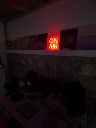 A dimly lit room featuring a white shelf displaying vinyl records and a brightly lit "ON AIR" sign. Adjacent to the shelf is a textured wall with a rocky surface. A desk is visible, cluttered with objects including a laptop, headphones, and other miscellaneous items. Overall the room is dark, making details difficult to discern.