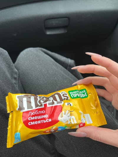 A close-up, eye-level shot shows a person's hand with long, light-pink acrylic nails holding a yellow M&M's candy bar wrapper. The wrapper features the M&M's logo in brown and white, a red oval with Russian text that reads "Люблю смешно смеяться" (I love to laugh funny), and a cartoon M&M character. A green banner on the wrapper says "ВЫИГРЫВАЙ ПРИЗЫ" (Win prizes). The lower left corner of the wrapper displays nutritional information: "В 1 ПОРЦИИ (45 г):" (per serving (45g):), "ККАЛ 225" (kcal 225), and "9%". The background consists of dark grey denim pants and a dark grey car interior. The image is cropped to focus on the candy and hand, with no discernible city or Russia context present within the frame.