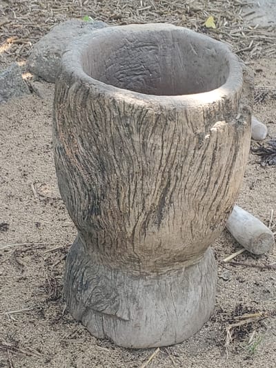 A close-up shot shows a weathered, hollowed-out wooden mortar resting on sandy ground. The mortar's exterior is rough and textured with visible grain patterns and vertical striations, indicating age and use. The opening at the top is circular and appears to be worn smooth. The surrounding environment consists of dry, light-colored sand mixed with small twigs and debris. In the background, a larger, rough-textured stone or wooden object is partially visible to the left of the mortar. Two cylindrical wooden pieces lie on the ground to the right and behind the mortar. The lighting suggests it is daytime, with a bright highlight visible inside the mortar. The scene is located in Dawaki, Nigeria.