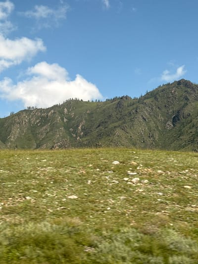 The image shows a mountainous landscape in Tuekta, Russia, under a blue sky with scattered white clouds. The foreground is a grassy field with rocks and sparse vegetation. The midground features a green, rocky mountain slope with scattered trees and shadows. The background is dominated by the mountain range and the sky. There are no people or animals visible in the image, and no discernible action is taking place. The scene appears to be a natural, undeveloped area.