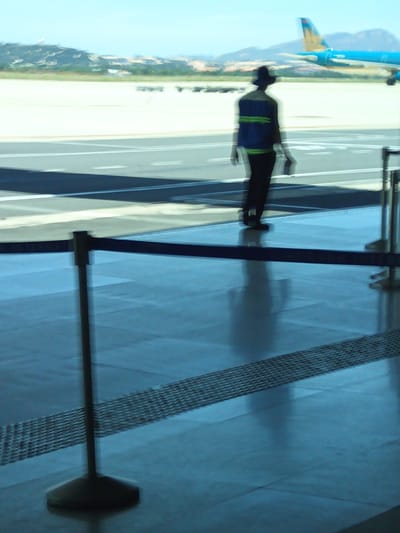 An out-of-focus, full-body shot captures a person wearing a dark hat, a dark blue vest with two horizontal green stripes, and dark pants, standing at what appears to be an airport in Cam Ranh, Vietnam. The individual is facing away from the camera, with their right hand slightly extended forward. Behind the person, a blue and yellow airplane is visible on the tarmac, with mountains in the background under a clear sky. The foreground shows a reflective blue floor with a dark barrier and a metal grate pattern.
