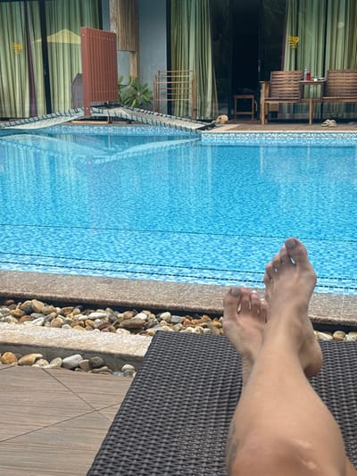 This image depicts an outdoor leisure scene in Ban Tha Muang, Thailand. In the foreground, a person's bare legs and feet, with light-colored toenail polish, are crossed at the ankles and rest on a dark brown woven synthetic lounge chair. The skin on the legs exhibits lighter patches and a faint tattoo is visible on the left calf. To the left of the lounge chair, wooden deck tiles are visible. Immediately in front of the person, a border of various natural river rocks separates the lounge area from the edge of a swimming pool.
The midground features a clear, rectangular swimming pool with bright blue mosaic tiles, reflecting the sky and surrounding structures. A dark blue tiled line marks the waterline.
In the background, beyond the pool, a building is visible with multiple large glass sliding doors covered by light green curtains. To the left, a light-colored, net-like hammock structure is positioned partially over the pool from a wooden base. A tall, brown slatted wooden screen or divider stands near the center of the background, with a small green plant behind it. To the right, two wooden slatted benches flank a small wooden side table holding a red can and another object. A pair of light-colored sandals rest on the wooden deck near the right bench. Further right, a wooden rack or drying stand is present. The overall environment suggests a resort or hotel setting.