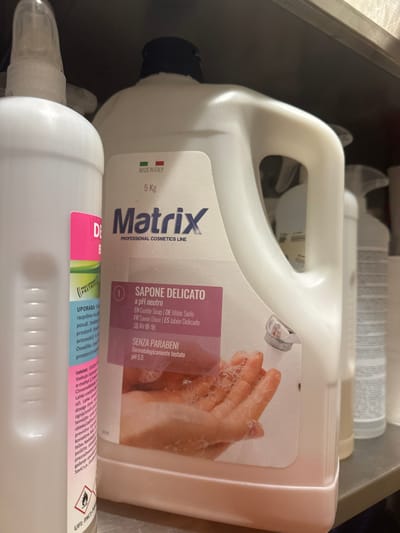 This image depicts a collection of commercial cleaning and cosmetic products stored on a light-colored shelf within a location in Radovljica, Slovenia. The central and most prominent object is a large white 5 kg plastic jug with a handle and a black screw-on cap, labeled "Matrix PROFESSIONAL COSMETICS LINE." Its main label, partially pink-purple, identifies the product as "SAPONE DELICATO a pH neutro" (Gentle Soap, pH 5.5, dermatologically tested, paraben-free), with translations including English, German, French, and Spanish. The label also features an illustration of hands with soap bubbles and indicates "MADE IN ITALY." The liquid inside the jug appears light pink. To the left, a tall, slender white plastic bottle with a white spray nozzle is partially visible. Its label, featuring pink and green sections, displays Slovenian text regarding usage ("UPORABA"), the product name "DEO," a UFI code ("PXCX-P0M9"), and a flame hazard symbol. To the right of the central jug, another white plastic container with a white spray nozzle is partially visible, followed by a clear plastic bottle containing a pale yellow liquid. Further right, rolls of white paper or towels are visible. A dark surface, likely another shelf or wall, is situated above and behind the items.