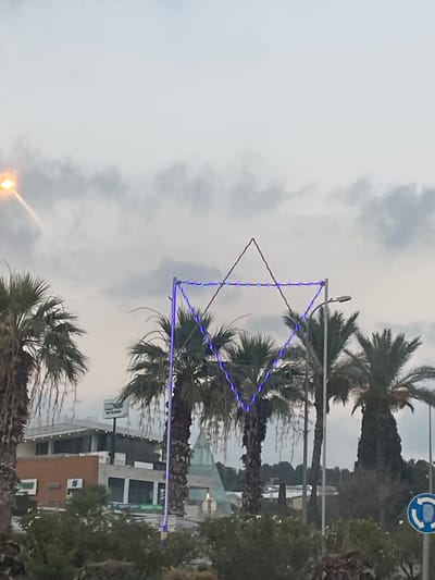 The image depicts a street scene in Shlomi, Israel, under a cloudy sky. A prominent Star of David decoration, outlined with blue lights, is suspended above several palm trees. In the background, buildings and foliage are visible, suggesting an urban or suburban setting. The lighting indicates it is either early morning or late afternoon. There are no people visible in the frame, and the focus is on the decorative element and the surrounding environment.