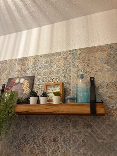 A wooden shelf is mounted on a wall covered with patterned tiles. On the shelf, from left to right, are a cascading green plant, a framed picture of cherry tomatoes and pasta, three small white planters with succulent plants, a small wooden clock, a blue glass bottle, and a small blue lidded container. The wall above the tiled area is a textured white surface with dappled shadows. The scene is illuminated by overhead lighting, casting shadows on the textured wall.