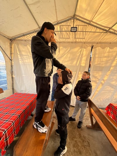 Location: Likely inside a tent in Zlatna Panega, Bulgaria, given the description.
Description: The image depicts four individuals within a tent-like structure. The tent's framework is visible, with a white or light-colored fabric forming the walls and ceiling. One person wearing a black cap and a black denim jacket appears to be gesturing towards their mouth. Another individual, dressed in a black hoodie with white text and black pants, is being touched on the head by the first person. A third person stands behind them, wearing a blue shirt. A fourth person is visible, wearing a black puffy jacket and grey pants. The people are positioned near wooden benches covered with red and white plaid fabric. The flooring appears to be concrete.
