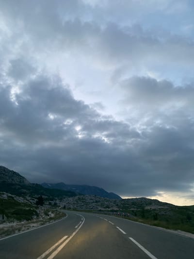 The image depicts a winding asphalt road with double white lines, leading through a mountainous landscape. The sky is overcast with dramatic, dark clouds, suggesting impending weather changes or a stormy atmosphere. Rocky outcrops and sparse vegetation, primarily grass and some trees, are visible on the hillsides flanking the road. In the distance, mountain peaks are shrouded in mist or shadow, adding to the dramatic and somewhat somber mood of the scene. The road appears to be empty of vehicles, and no people are visible. The lighting suggests it is either early morning or late evening, possibly twilight. The location is identified as Pašina voda, Montenegro.