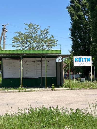 The image shows a roadside view in Staryi Saltiv, Ukraine. A small, green-roofed structure with closed, bullet-ridden metal shutters stands adjacent to a narrow, worn asphalt road. Overgrown grass and weeds line the road's edge. A sign with the word "Квіти" (Flowers) in blue Cyrillic letters is visible above the structure. A utility pole with power lines is seen in the background, and trees are scattered throughout the scene, with a blue sky visible above. The overall scene appears to be in a state of neglect, with evidence of damage and disrepair on the building.